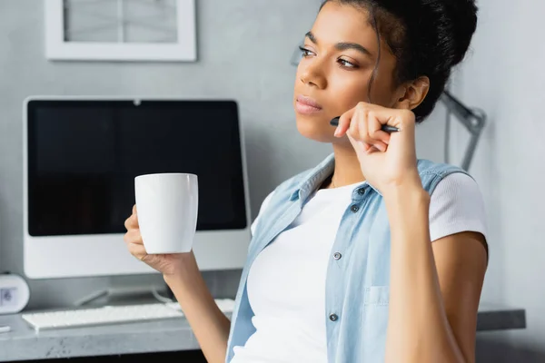 Reflexivo freelancer afroamericano sosteniendo taza de té y pluma cerca del monitor de la computadora sobre fondo borroso - foto de stock