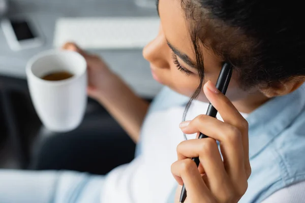 Overhead view of african american freelancer holding pen and cup of tea, blurred background — Stock Photo