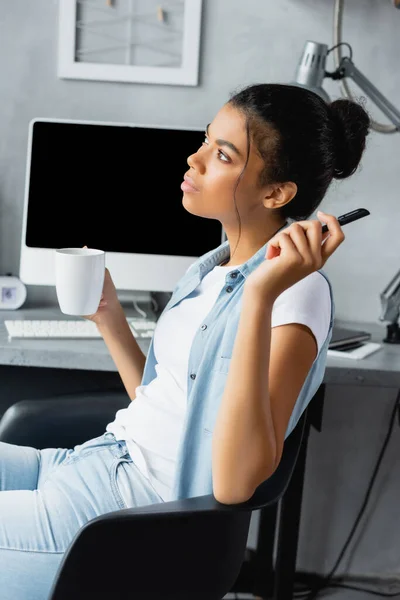 Pensoso afroamericano freelance che tiene penna e tazza mentre seduto vicino al monitor del computer — Foto stock