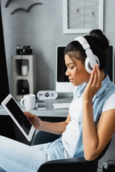 Visão lateral do freelancer afro-americano em fones de ouvido sem fio segurando tablet digital com tela em branco, fundo borrado — Fotografia de Stock