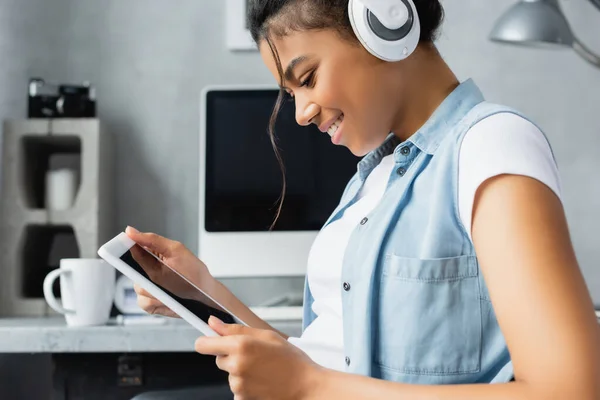 Side view of smiling african american freelancer in wireless headphones using digital tablet with blank screen, blurred background — Stock Photo