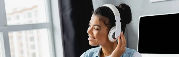 Femme freelance africaine joyeuse écoutant de la musique dans des écouteurs sans fil à la maison, bannière — Photo de stock