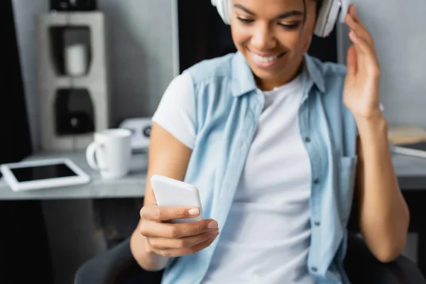 Allegro freelance afroamericano che tiene lo smartphone mentre ascolta musica in cuffie wireless, sfondo sfocato — Foto stock
