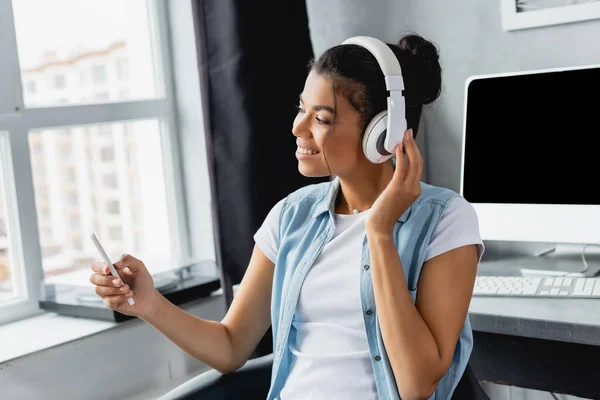 Heureux afro-américain freelance messagerie sur smartphone tout en écoutant de la musique dans les écouteurs sans fil à la maison — Photo de stock