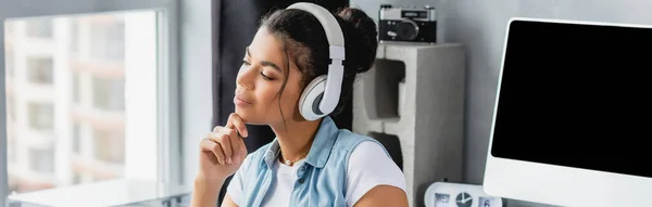 Smiling, dreamy african american freelancer listening music in wireless headphones near monitor, banner — Stock Photo