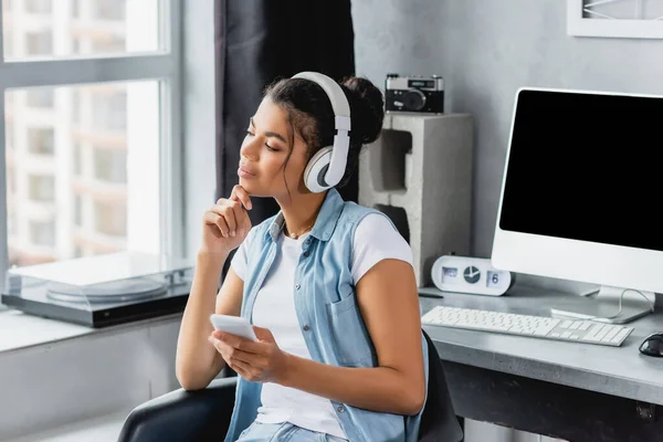 Dreamy african american freelancer in wireless headphones holding smartphone while sitting near computer monitor — Stock Photo