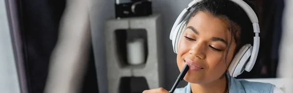 Femme afro-américaine souriante et rêveuse tenant un stylo tout en écoutant de la musique dans des écouteurs sans fil, au premier plan flou, bannière — Photo de stock