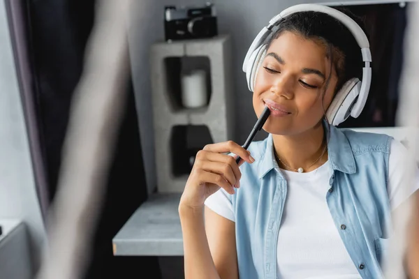 Smiling, dreamy african american woman holding pen while listening music in wireless headphones, blurred foreground — Stock Photo