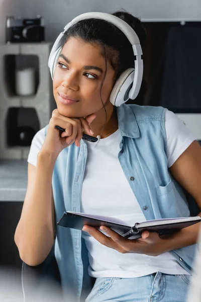 Nachdenklicher afrikanisch-amerikanischer Freiberufler mit drahtlosen Kopfhörern, der wegschaut, während er zu Hause sein Notizbuch hält — Stockfoto