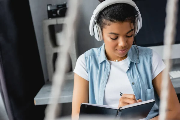 Young african american freelancer in wireless headphones writing in notebook on blurred foreground — Stock Photo