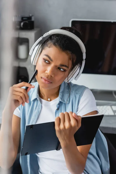 Freelancer afro-americano sério e atencioso em fones de ouvido sem fio segurando notebook e caneta, primeiro plano desfocado — Fotografia de Stock