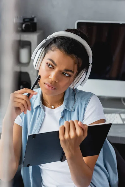 Serio, reflexivo afroamericano freelancer en auriculares inalámbricos que sostienen el cuaderno y la pluma, borrosa primer plano - foto de stock