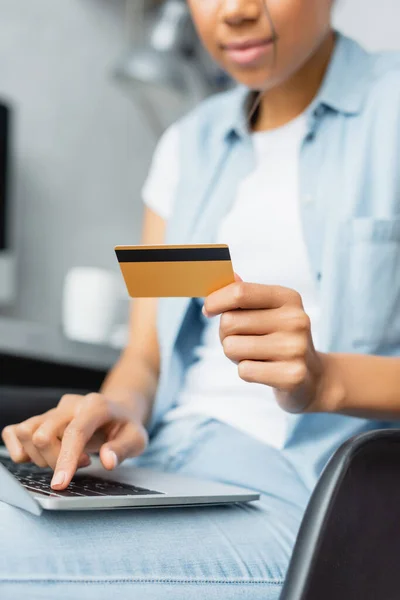 Vista parcial de la mujer afroamericana que sostiene la tarjeta de crédito mientras usa el ordenador portátil, borrosa primer plano - foto de stock