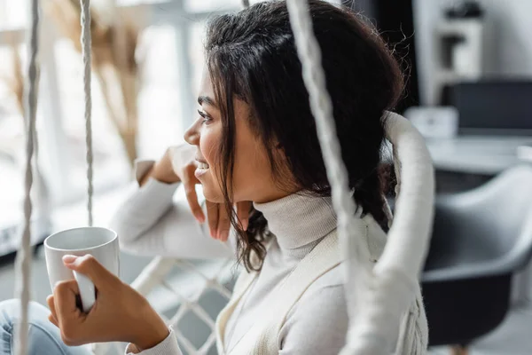 Happy african american woman relaxing in hanging chair with cup of tea and looking away, blurred foreground — Stock Photo