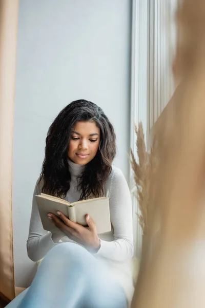 Young african american woman reading book near window at home, blurred foreground — Stock Photo
