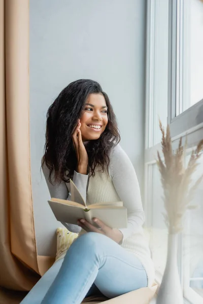 Joyeuse femme afro-américaine regardant par la fenêtre tout en étant assis sur le rebord de la fenêtre avec le livre — Photo de stock