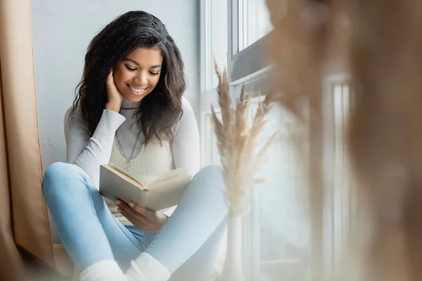 Sonriente mujer afroamericana leyendo libro cerca de ventana en casa en primer plano borroso - foto de stock