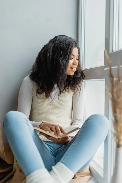 Smiling african american woman holding book while sitting on windowsill, blurred foreground — Stock Photo