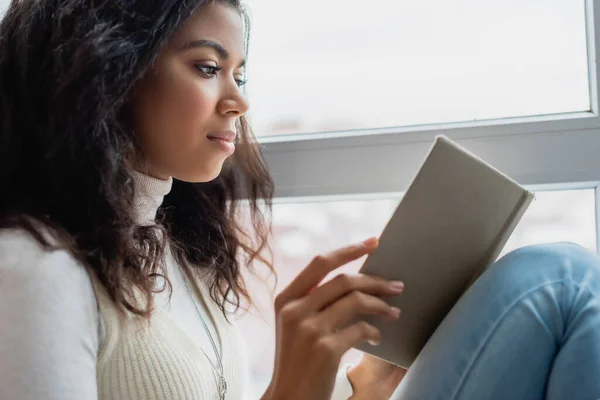 Молода афроамериканка читає книгу біля вікна вдома — стокове фото
