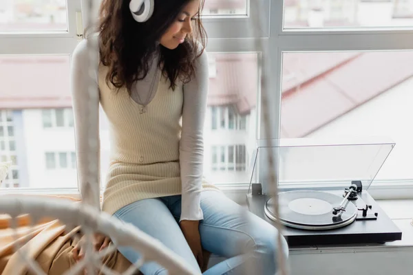 Joven mujer afroamericana en auriculares inalámbricos sentado cerca de tocadiscos en el alféizar de la ventana, borrosa primer plano - foto de stock