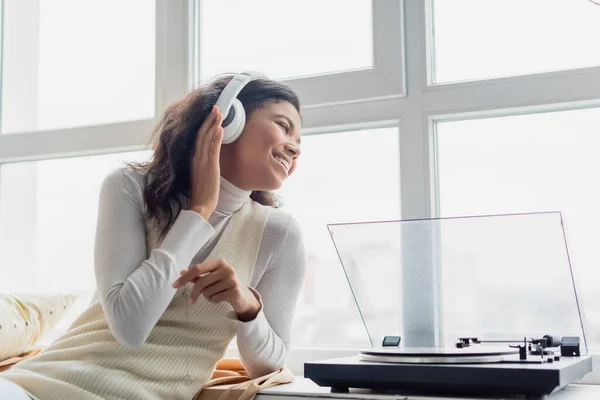 Mujer afroamericana feliz en auriculares inalámbricos mirando a través de la ventana cerca del reproductor de discos - foto de stock