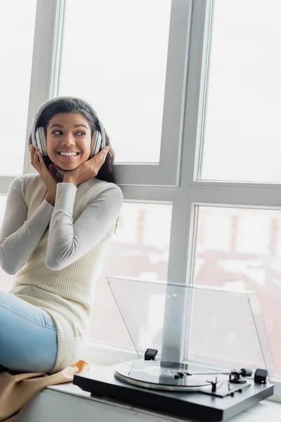 Fröhliche afrikanisch-amerikanische Frau berührt drahtlose Kopfhörer, während sie Musik in der Nähe des Plattenspielers auf der Fensterbank hört — Stockfoto