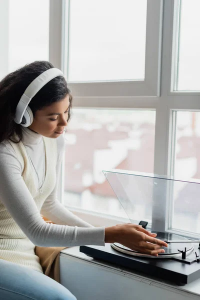 Young african american woman in wireless headphones turning on record player near window — Stock Photo