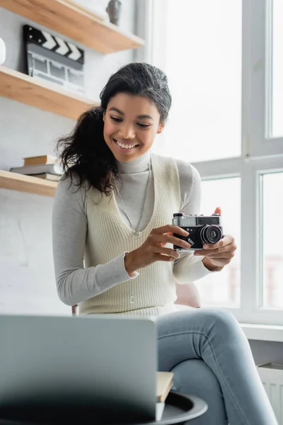 Mulher americana africana alegre segurando câmera retro enquanto olha para o laptop em primeiro plano borrado — Fotografia de Stock