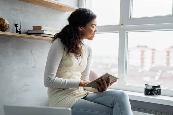 Sonriente mujer afroamericana sosteniendo libro mientras está sentado cerca de la cámara de fotos vintage en el alféizar de la ventana - foto de stock