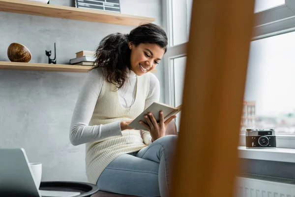 Fröhliche afrikanisch-amerikanische Frau liest Buch, während sie neben Vintage-Kamera auf Fensterbank sitzt, verschwommener Vordergrund — Stockfoto