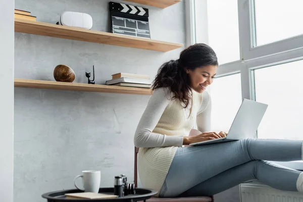 Happy african american woman typing on laptop near cup of tea and vintage camera on coffee table, blurred foreground — Stock Photo