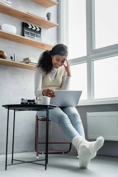 Fröhliche afrikanisch-amerikanische Frau blickt auf Laptop neben Teetasse und Vintage-Kamera auf Couchtisch — Stockfoto