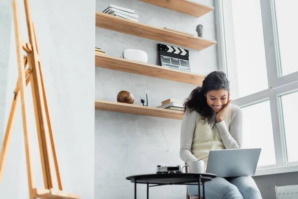 Sorridente donna africana americana che utilizza il computer portatile a casa vicino cavalletto in primo piano sfocato — Foto stock