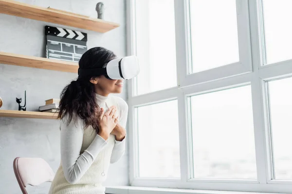 Donna afro-americana sorridente in auricolare vr che si tiene per mano sul petto vicino alla finestra — Foto stock