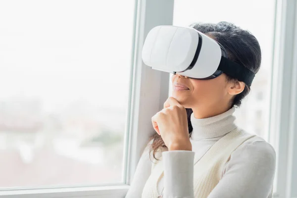 Lächelnde afrikanisch-amerikanische Frau berührt Kinn, während sie ihr Headset in der Nähe des Fensters benutzt — Stockfoto