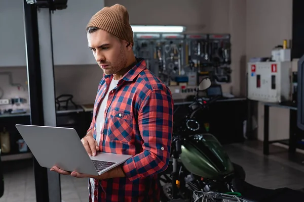 Giovane meccanico in camicia a quadri e berretto utilizzando laptop in officina vicino moto su sfondo sfocato — Foto stock