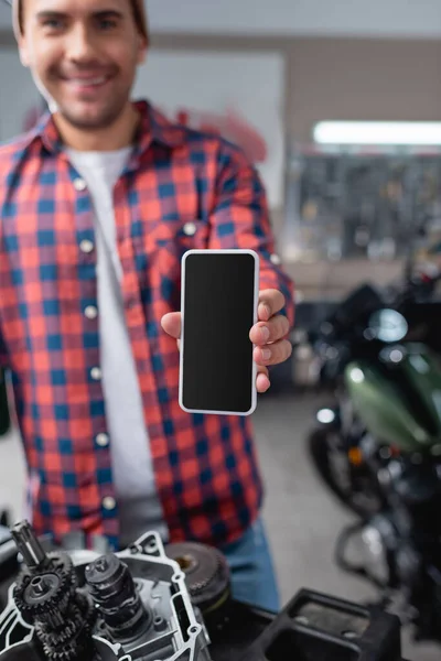 Lächelnder Mechaniker zeigt Smartphone mit leerem Bildschirm in der Nähe von Motorrad-Ersatzteilen, verschwommener Hintergrund — Stockfoto