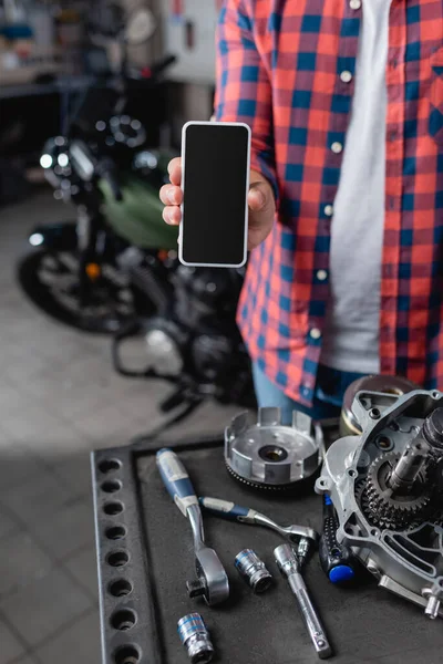 Vista parcial del mecánico que muestra el teléfono móvil con la pantalla en blanco cerca de piezas de repuesto y llaves de motocicleta, fondo borroso - foto de stock