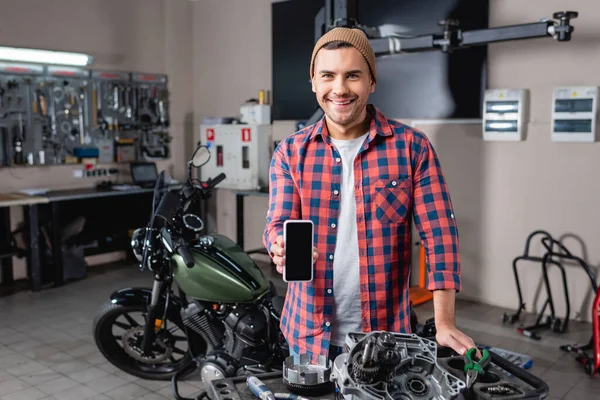 Mécanicien joyeux montrant téléphone portable avec écran blanc près de pièces de rechange et moto en atelier — Photo de stock