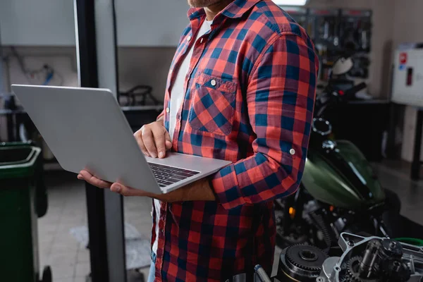 Cropped view of technician in plaid shirt with laptop near motorcycle spare parts in garage — Stock Photo