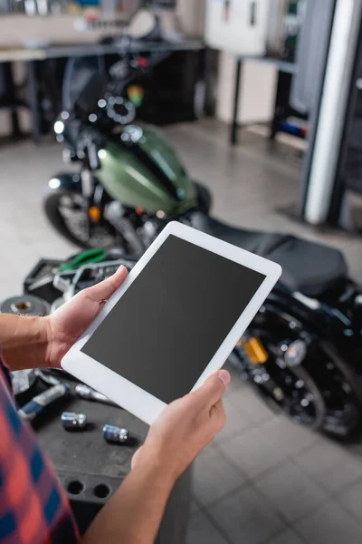 Обрізаний вид ремонстранта, що тримає цифровий планшет з порожнім екраном біля мотоцикла на розмитому фоні — стокове фото