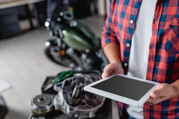 Vista recortada del mecánico que sostiene la tableta digital con la pantalla en blanco cerca de piezas de repuesto de la motocicleta en fondo borroso - foto de stock