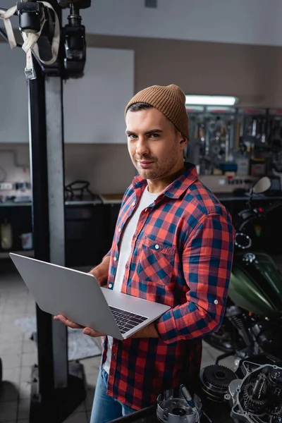 Young technician in checkered shirt and beanie smiling at camera while standing with laptop in garage — Stock Photo