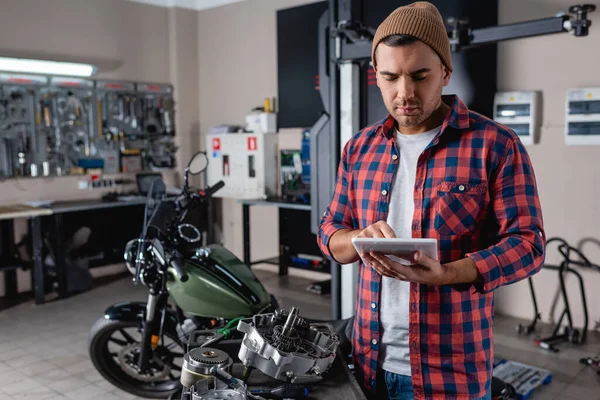 Joven mecánico en gorro y camisa a cuadros usando tableta digital cerca de piezas de repuesto y motocicleta sobre fondo borroso - foto de stock