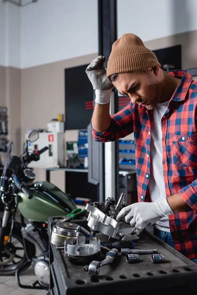 Young mechanic touching head while checking disassembled motorcycle gearbox — Stock Photo