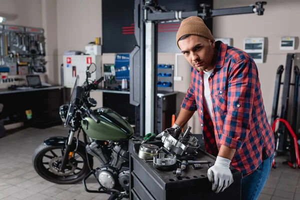 Junger Mechaniker in kariertem Hemd und Mütze blickt in die Kamera neben zerlegtem Motorradgetriebe — Stockfoto