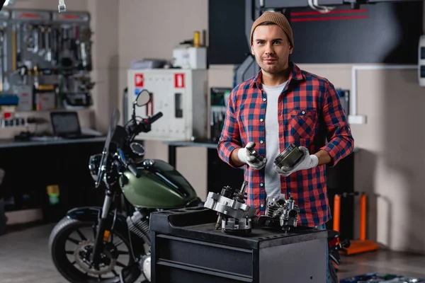 Lächelnder Mechaniker in kariertem Hemd und Handschuhen, der Zahnräder neben zerlegtem Motorradgetriebe hält — Stockfoto