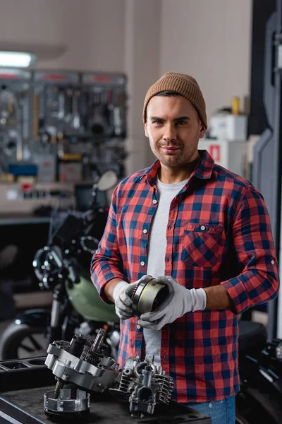 Jovem mecânico em camisa quadriculada e gorro olhando para a câmera perto desmontada motocicleta caixa de velocidades — Fotografia de Stock