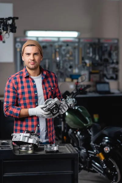 Young mechanic in plaid shirt and beanie looking at camera while holding part of gearbox near motorbike on blurred background — Stock Photo