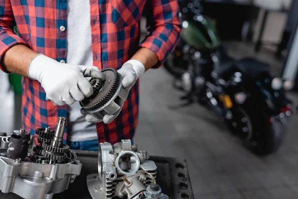 Ausgeschnittene Ansicht des Mechanikers in kariertem Hemd und Handschuhen, der das Zahnrad in der Nähe des zerlegten Motorradgetriebes hält — Stockfoto